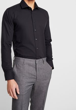 Hombre vistiendo una camisa negra ajustada de botones metida en pantalones de vestir grises con textura, con las manos en los bolsillos, frente a un fondo claro y liso.