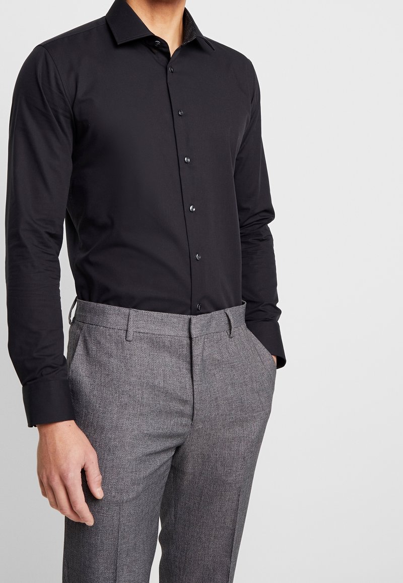 Man som bär en åtsittande svart skjorta med knappar instoppad i grå texturerade kostymbyxor med händerna i fickorna, mot en enkel ljus bakgrund.