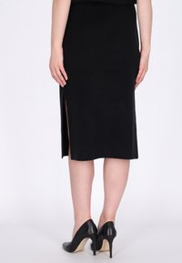 Eine Frau trägt einen schwarzen Midirock mit einem Seitenschlitz und schwarze High Heels, die vor einem schlichten hellen Hintergrund steht.