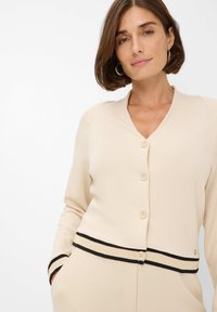 Vrouw met kort bruin haar, gekleed in een crèmekleurige cardigan met knopen en zwarte gestreepte boorden en onderrand, poseert met handen in de zakken tegen een witte achtergrond.