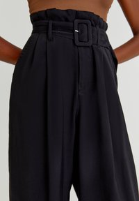 Pantalons noirs taille haute avec une large ceinture en tissu et boucle rectangulaire, portés avec un haut sans manches marron, les mains dans les poches.