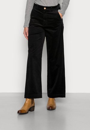 Personne portant un pantalon large en velours côtelé noir, un haut gris à manches longues et des bottines marron, debout sur un sol gris.