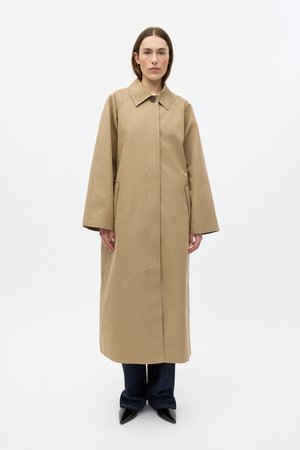 Abrigo beige hasta la rodilla con cuello, cierre frontal con botones, bolsillos laterales y mangas anchas. Llevado sobre vaqueros azul oscuro y zapatos negros puntiagudos.