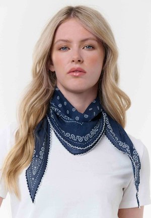 Blonde woman with loose hair wearing a white top and a navy blue patterned bandana tied around her neck.