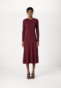 Burgundy bordázott midi ruha hosszú ujjal, gombos elülső résszel és két mellzsebbel. Fekete gombokkal díszített, fekete platform szandálokkal viselve.