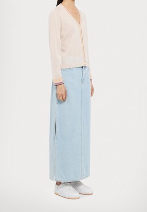 Kobieta ubrana w jasnobeżowy rozpinany sweter, długą jasnoniebieską spódnicę jeansową z bocznym rozcięciem oraz białe trampki, stojąca bokiem.