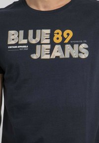 T-shirt en coton bleu marine avec un imprimé graphique en blanc et jaune, comportant le texte "BLUE 89" et "VINTAGE APPARELS EST. 1989".