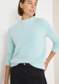 Maglione a collo alto di un blu chiaro, lavorato a maglia con un motivo testurizzato, bordo e polsini a costine. Abbinato a jeans neri. Leggera morbidezza evidente nel tessuto.