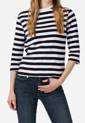 Femme aux longs cheveux portant un haut à manches longues rayé noir et blanc et un jean bleu foncé, une main dans la poche et l’autre sur la hanche.