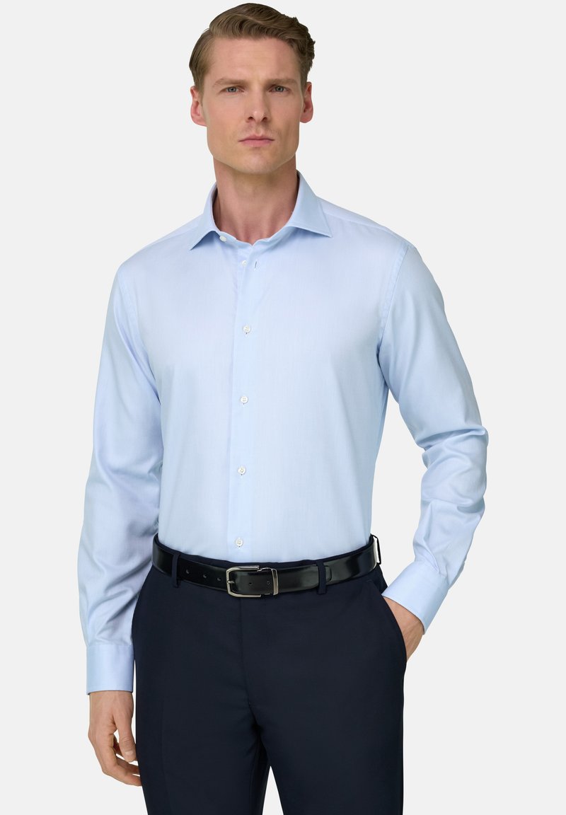 Homme portant une chemise habillée à manches longues bleu clair rentrée dans un pantalon sombre avec une ceinture noire, debout avec une main dans la poche.