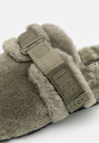 Olivgrön toffel med fluffig shearling-textur, justerbar rem med UGG-logotyp och en svart gummisula.