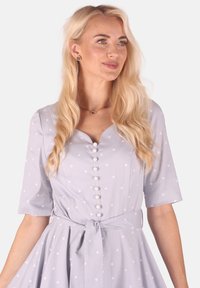 Helles blaues Kleid mit weißen Punkten, kurzen Ärmeln, V-Ausschnitt und Knopfleiste. Mit einem taillierten Gürtel und einem ausgestellten Rock.