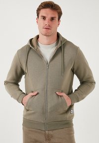 Sudadera con cremallera en verde oliva, con capucha ajustable, bolsillos frontales, puños acanalados y dobladillo; confeccionada en una suave tela texturizada.