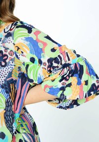 Arm bøjet med hånden på hoften iført en multicolor abstrakt mønstret bluse med puffede ærmer mod en hvid baggrund.