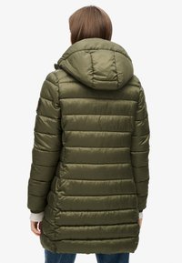 Abrigo acolchado verde olivo con capucha, que presenta secciones acolchadas horizontales y un parche con el logo en la manga izquierda. Textura suave y brillante.