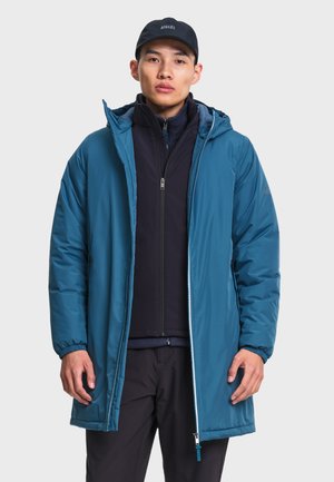 Jeune homme portant une veste d'hiver bleue à capuche sur un pull zippé noir et une casquette noire, debout devant un fond clair uni.