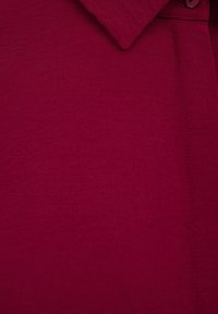 Burgundská košile s texturovaným povrchem, s knoflíkovým límcem a viditelnými švy. Hladká tkanina s jemným leskem.