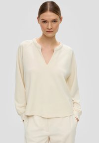 Lichtbeige langemouwtop met een V-hals en raglanmouwen. Zacht materiaal en een relaxte pasvorm, gecombineerd met lichte geribde broek.