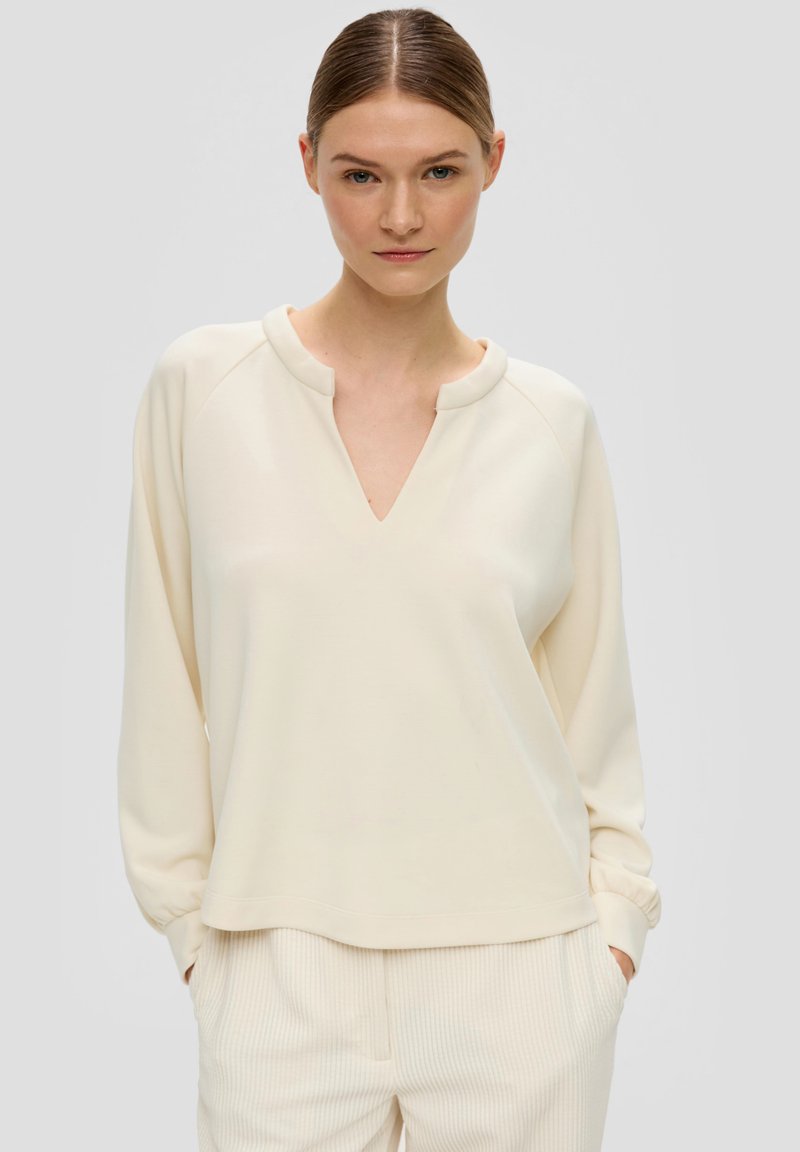 Lichtbeige langemouwtop met een V-hals en raglanmouwen. Zacht materiaal en een relaxte pasvorm, gecombineerd met lichte geribde broek.