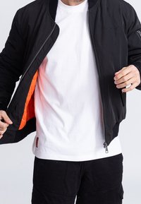 Homme en train de fermer une veste noire révélant une doublure orange, portant un t-shirt blanc et un pantalon noir, cadré au niveau du cou et des cuisses.