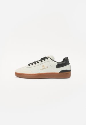 White leather sneaker with black accents and logo. Features round toe, lace-up front, and rubber sole with a textured brown bottom.