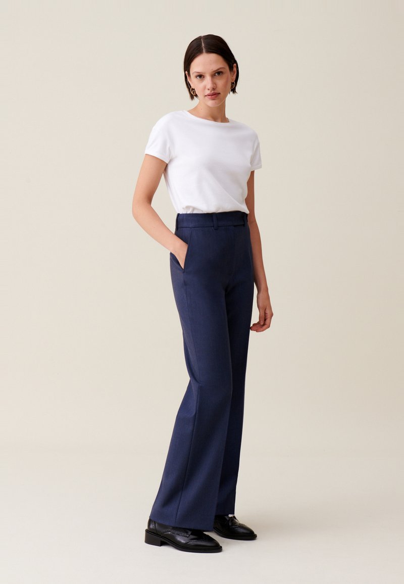 Marineblau zugeschnittene Hose mit geradem Bein, kombiniert mit einem weißen T-Shirt mit kurzen Ärmeln. Dunkle Schuhe vervollständigen den Look. Glatte Stofftextur.
