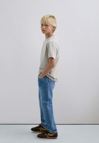 T-shirt gris clair à manches courtes, jeans en denim bleus, baskets marron avec des accents jaunes. Le modèle se tient de côté avec les mains dans les poches.