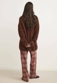 Frau mit langen braunen Haaren steht vor einer Wand, trägt einen braunen flauschigen Pullover und rosa karierte Hose mit dunklen Hausschuhen.