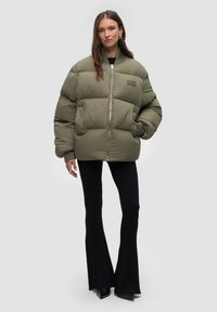 Olive green puffer jacket with a zip front and ribbed cuffs. Worn over a black ribbed outfit, featuring a logo patch on the chest.