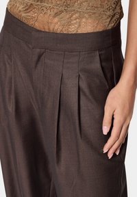 Pantalons plissés marron avec une finition textile lisse. Comprend une taille large et une texture subtile. Main placée sur le côté.