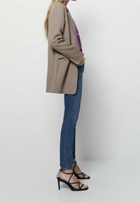 Beige taillierter Blazer, figurbetonte blaue Jeans und schwarze Riemchen-High-Heels. Der Stil ist sauber, ohne sichtbare Muster oder Accessoires.