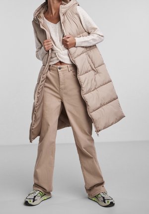 Beżowa kamizelka puchowa z kapturem, zestawiona z jasnym swetrem i jasnobrązowymi spodniami typu wide-leg. Buty mają szaro-biało-neonowo zielony design.