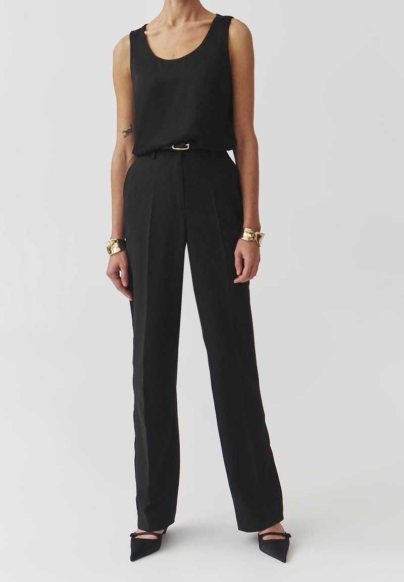Personne portant un haut noir sans manches, un pantalon noir taille haute avec ceinture, des chaussures noires à bout pointu et des bracelets en or aux deux poignets.