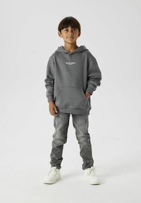 Grijze hoodie met een voorzak en witte tekst "Studio editie." Gecombineerd met vervaagde grijze jeans en witte sneakers. Effen achtergrond.