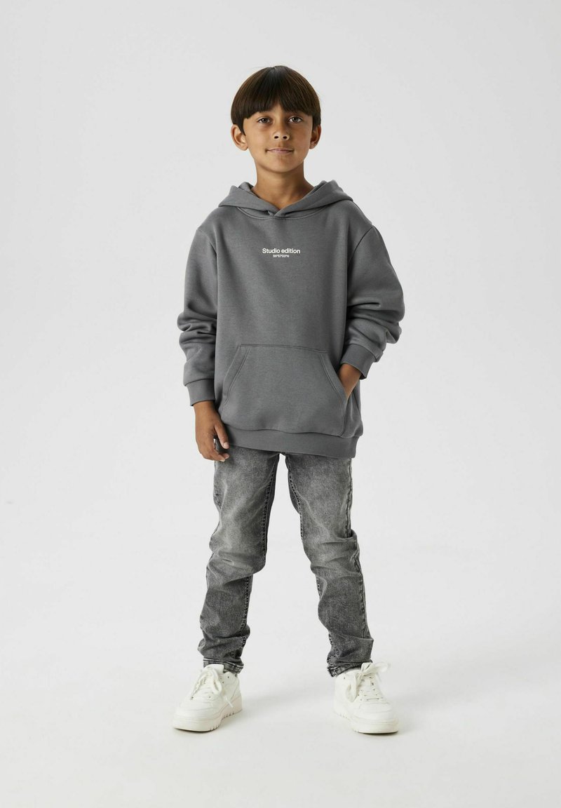 Grijze hoodie met een voorzak en witte tekst "Studio editie." Gecombineerd met vervaagde grijze jeans en witte sneakers. Effen achtergrond.