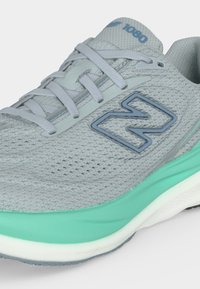 Chaussure de sport gris clair avec tige en mesh, semelle intermédiaire turquoise, lacets blancs et logo "N" gris foncé sur le côté.