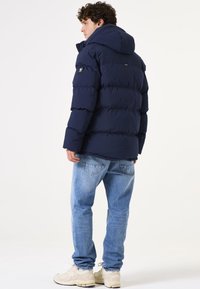 Marineblauwe pufferjack met een capuchon, gequilt ontwerp en een logo-opdruk op de mouw. Draag het over lichtblauwe jeans en sneakers.