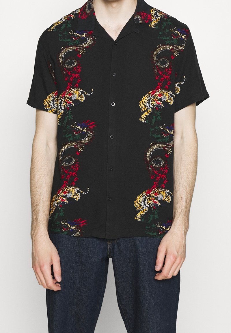 Chemise noire à manches courtes boutonnée avec des imprimés colorés de dragon et de tigre portée par une personne debout, les bras détendus.