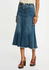 Gonna midi in denim blu scuro, design svasato con orlo grezzo, tasche frontali e cuciture a contrasto. Indossata con sandali con tacco marroni.
