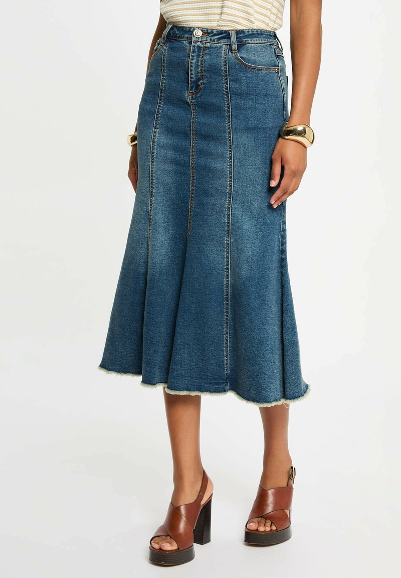Gonna midi in denim blu scuro, design svasato con orlo grezzo, tasche frontali e cuciture a contrasto. Indossata con sandali con tacco marroni.