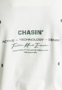 Tessuto bianco con testo grafico nero. Presenta il nome del marchio "CHASIN'" e le frasi "ACTIVE - TECHNOLOGY - DENIM" in diversi stili di carattere.
