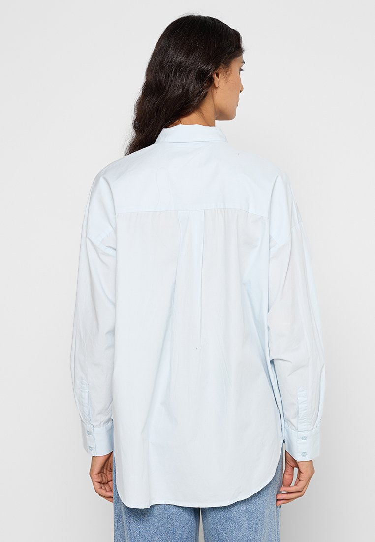 Camicia oversized azzurro chiaro con maniche lunghe e polsini con bottone, dotata di spalline posteriori e orlo dritto. Tessuto di cotone morbido.