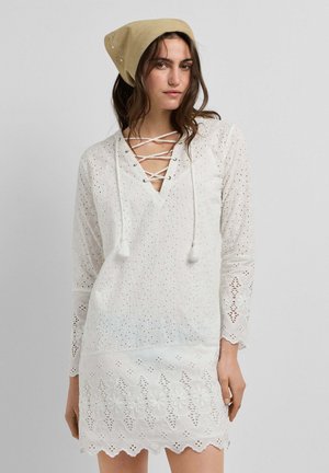 Vrouw draagt een witte oogjes kanten jurk met lange mouwen, gegolfde zoom, halslijn met kruislingse linten en een beige hoofddoek, staand tegen een grijze achtergrond.