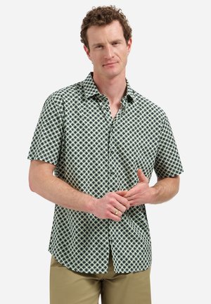 Homme portant une chemise à manches courtes à boutons avec un motif en losange noir et blanc, un pantalon beige, et une bague à la main gauche, debout devant un fond blanc.