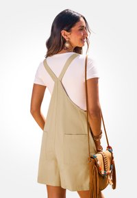 Beige stoffen overall met brede bandjes en twee zakken, gedragen over een wit shirt. Bruine schoudertas met kleurrijke accenten en franjes.