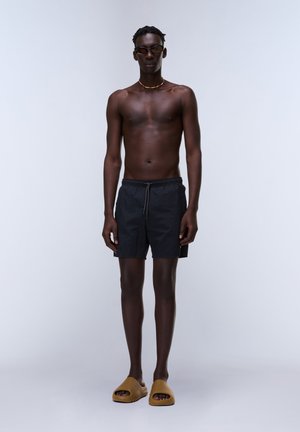 Großer dunkelhäutiger Mann, der schwarze Badeshorts, braune Slides, eine Sonnenbrille und eine Perlenkette trägt, steht vor einem schlichten Hintergrund.