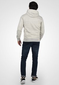 Lichtgrijze hoodie met textuurstof, geribde manchetten en onderrand, gecombineerd met donkerblauwe jeans en zwarte sneakers. Zicht van achter.