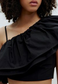 Schwarzes Crop-Top mit einem Schulterträger, das auf einer Seite ein großes Rüschendesign aufweist. Glatte Textur ohne sichtbare Muster oder Details.