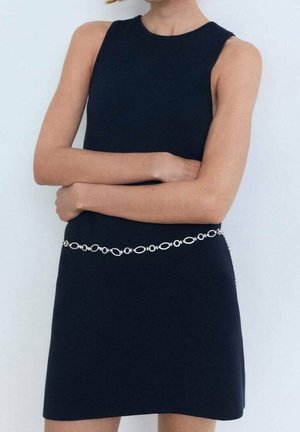 Ärmelloses schwarzes Kleid mit rundem Ausschnitt, accessorisiert mit einem silbernen Kettengürtel, Arme vor der Taille verschränkt.