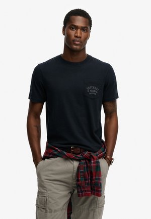 Granatowy bawełniany T-shirt z kieszenią na piersi ozdobioną fakturowanym logo. Noszony z beżowymi szortami typu cargo oraz czerwoną koszulą w kratę zawiązaną wokół talii.